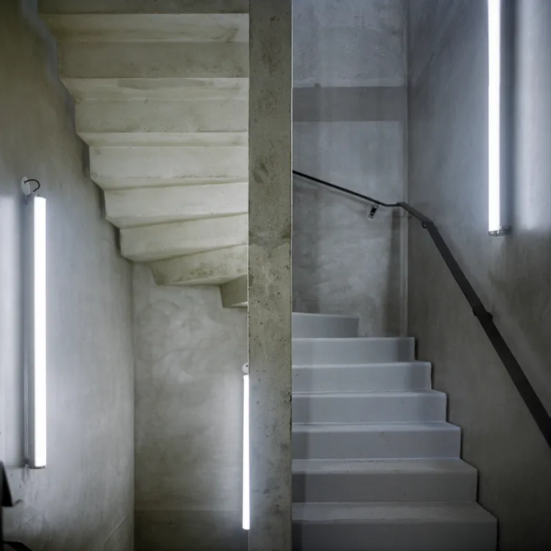 Concrete stairwell with vertical tube lighting and black metal handrail along gray plastered walls