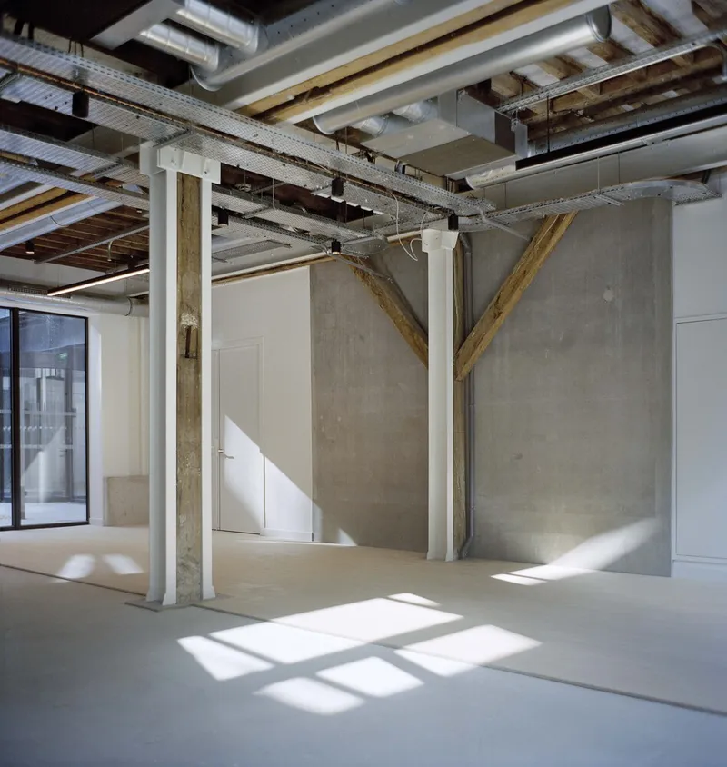 Open interior with exposed ceiling joists, diagonal timber bracing and sunlight casting window shadows on concrete