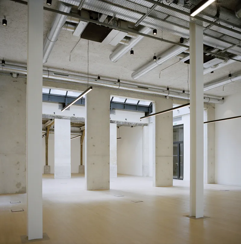 Open interior space with exposed concrete columns and ceiling ducts beneath a perforated metal screen skylight
