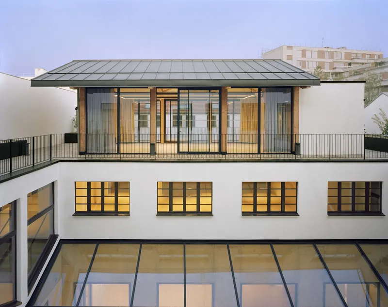 Courtyard view showing white rendered walls, glazed roof, metal-framed rooftop pavilion with timber screens at dusk