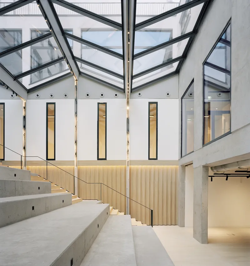 Interior atrium with angled steel-framed skylight, concrete seating tiers, and timber-clad wall below vertical slot windows