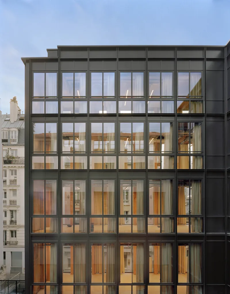 Gridded glass facade revealing timber-framed floors and interiors under an overcast sky