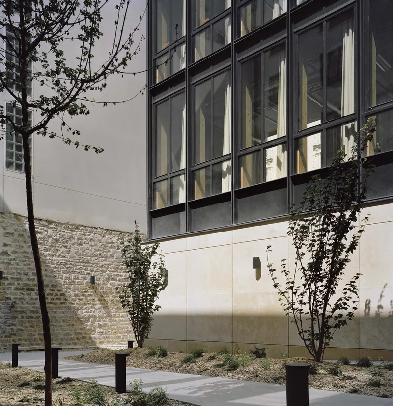 Gridded glass facade rising above stone base wall with young trees and climbing vines in the forecourt