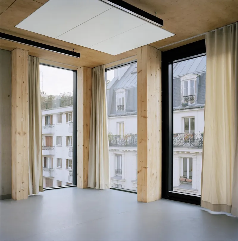 Interior corner with timber columns and exposed ceiling beams framing tall windows overlooking neighboring facades
