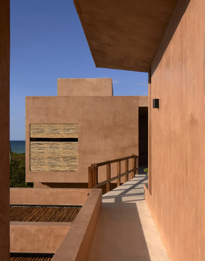 Open-air walkway with timber handrail connecting rammed earth walls under deep soffit overhang