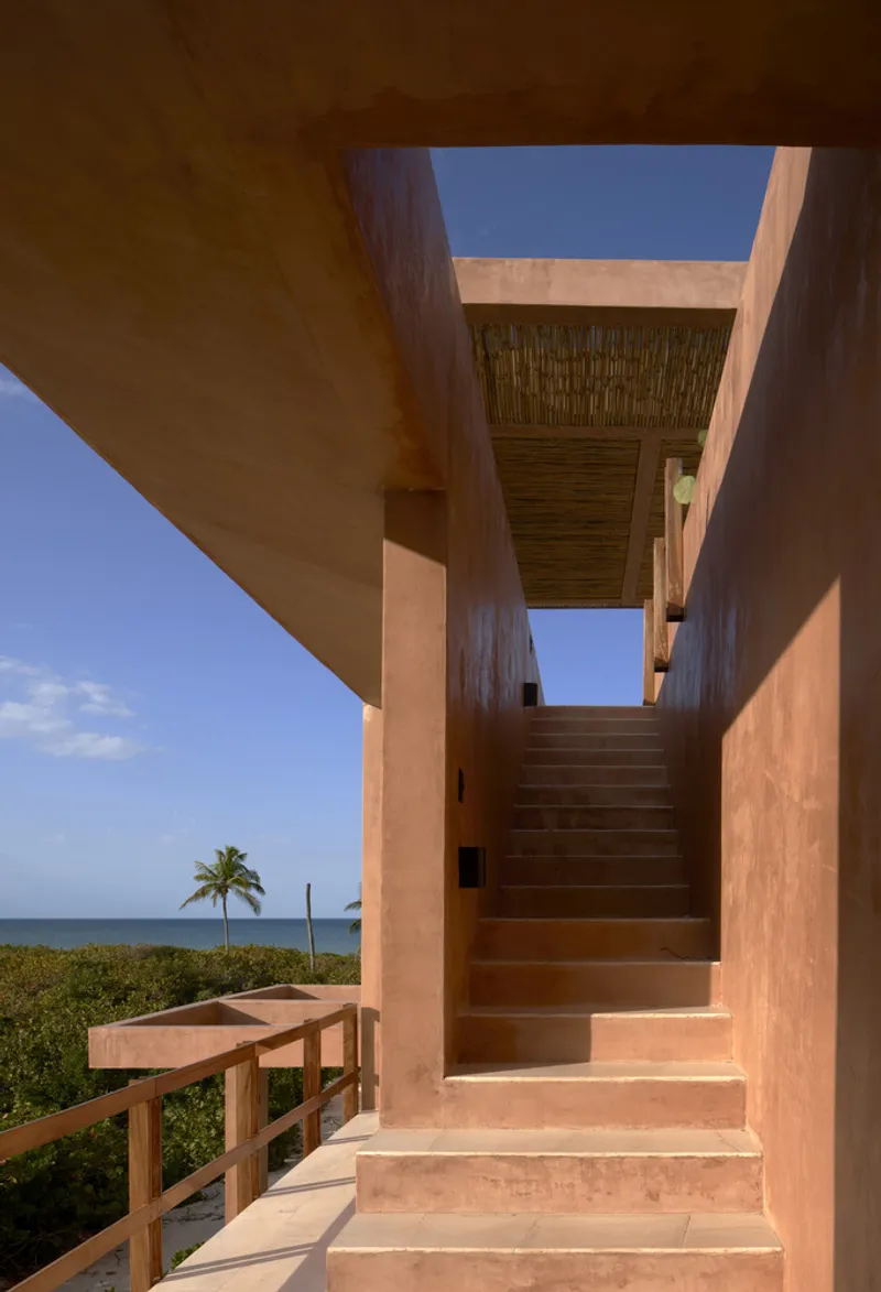 Rammed earth staircase rising between thick walls under a woven reed ceiling with palm trees and ocean beyond