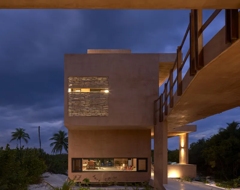Stacked concrete volumes with illuminated openings and timber bridge at dusk beside coconut palms