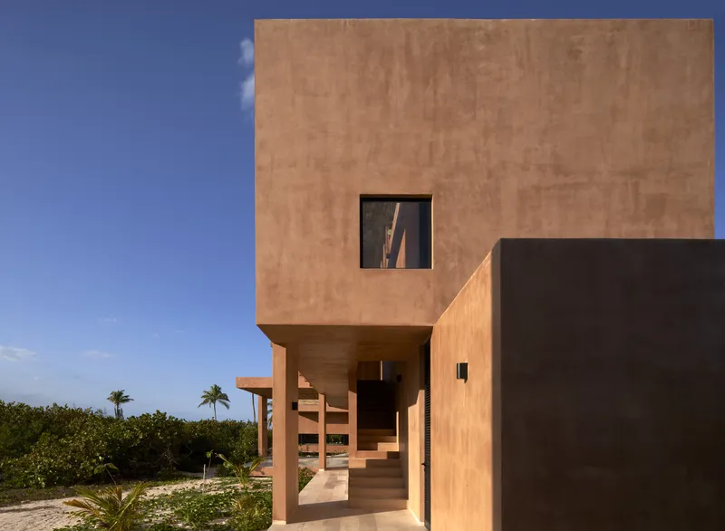 Cantilevered concrete box with square window above a covered staircase framed by coastal vegetation