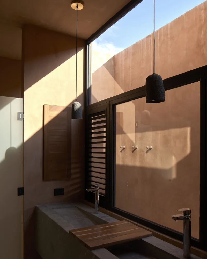 Bathroom with concrete trough sink and louvered window casting angular shadows in afternoon sunlight