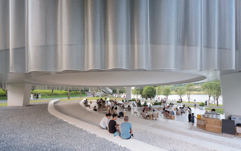 Covered public plaza with curved seating and perforated metal facade above people gathering on terrazzo flooring