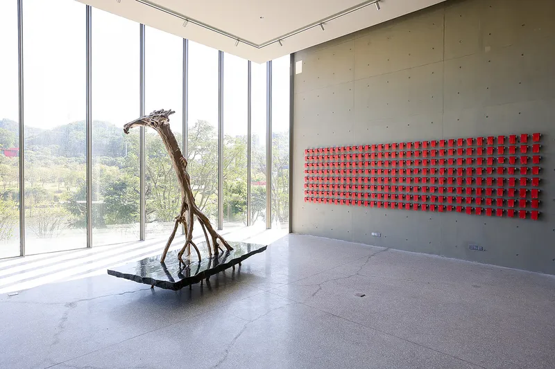 Corner gallery with full-height glazing, exposed concrete wall with red grid installation and bronze sculpture on floor