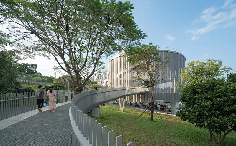 Elevated walkway with white metal railings curving past a cylindrical volume framed by mature trees