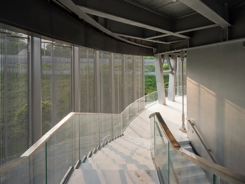 Curved staircase descending under an exposed black ceiling with mesh screens filtering daylight