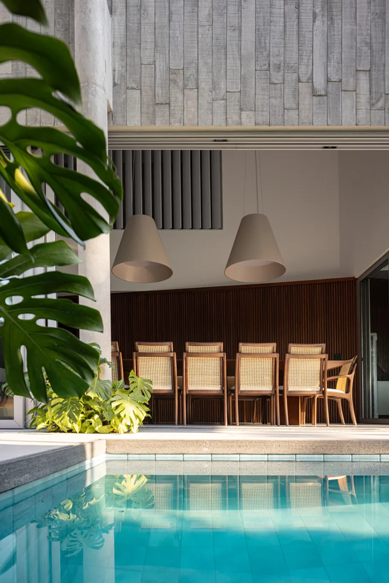 Pool edge reflecting the double-height space with slatted timber wall and cane-back chairs in shadow