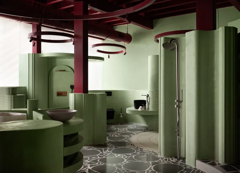 Bathroom interior with pale green partitions, curved walls, and patterned terrazzo flooring beneath exposed red ceiling beams
