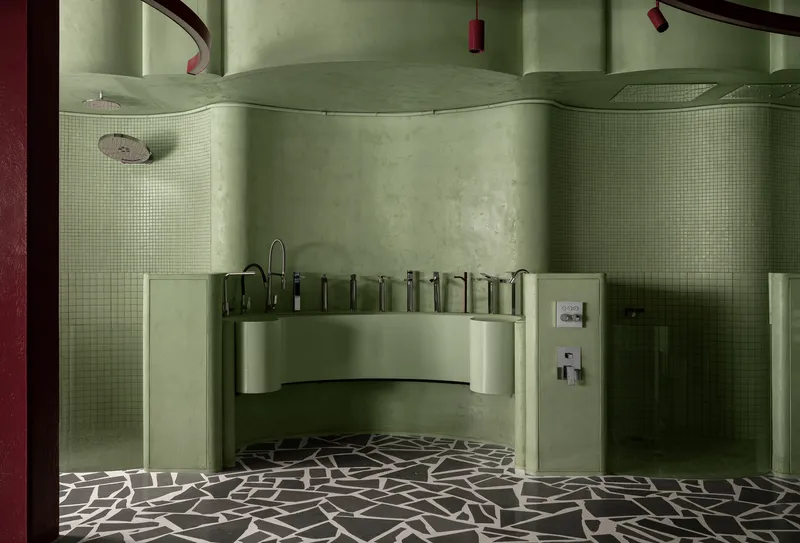 Curved vanity counter with multiple faucets flanked by tile-clad shower enclosures and terrazzo flooring