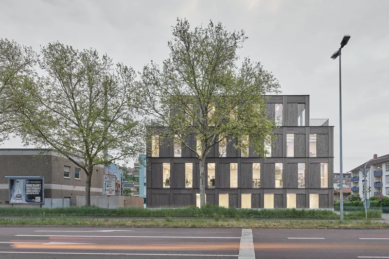 Street view of the timber-clad facade with vertical windows framed by bare trees at dusk