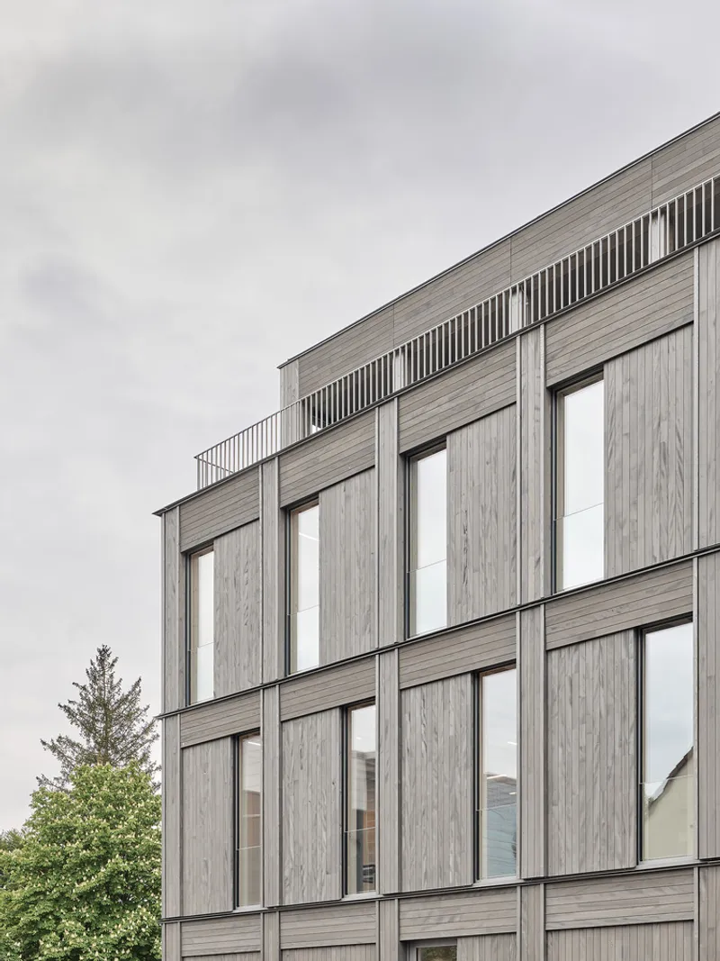 Upper facade detail of weathered timber cladding with tall vertical windows under an overcast sky