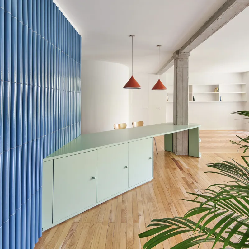 Dining area with pale green kitchen island, blue corrugated partition, exposed concrete beam, and red pendant lights