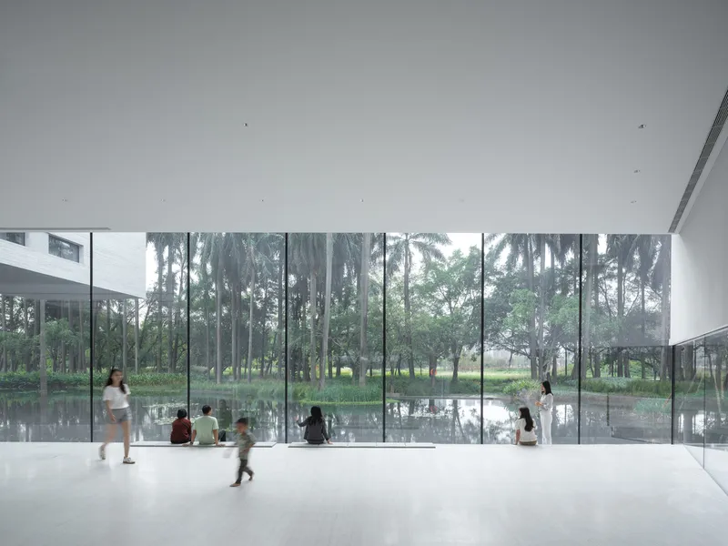 Floor-to-ceiling glass wall framing palm trees and pond with visitors seated and walking inside