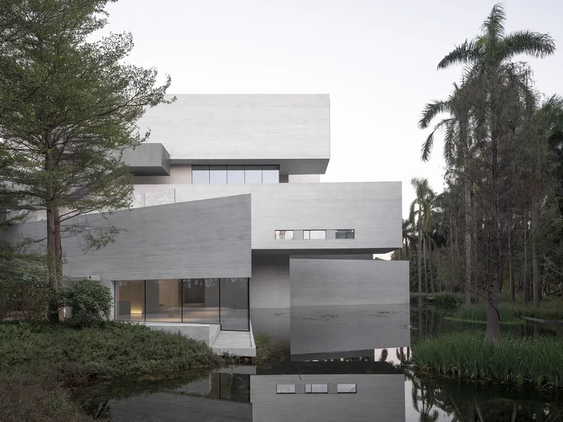 Cantilevered concrete facade with punched windows reflecting in the pond at dusk