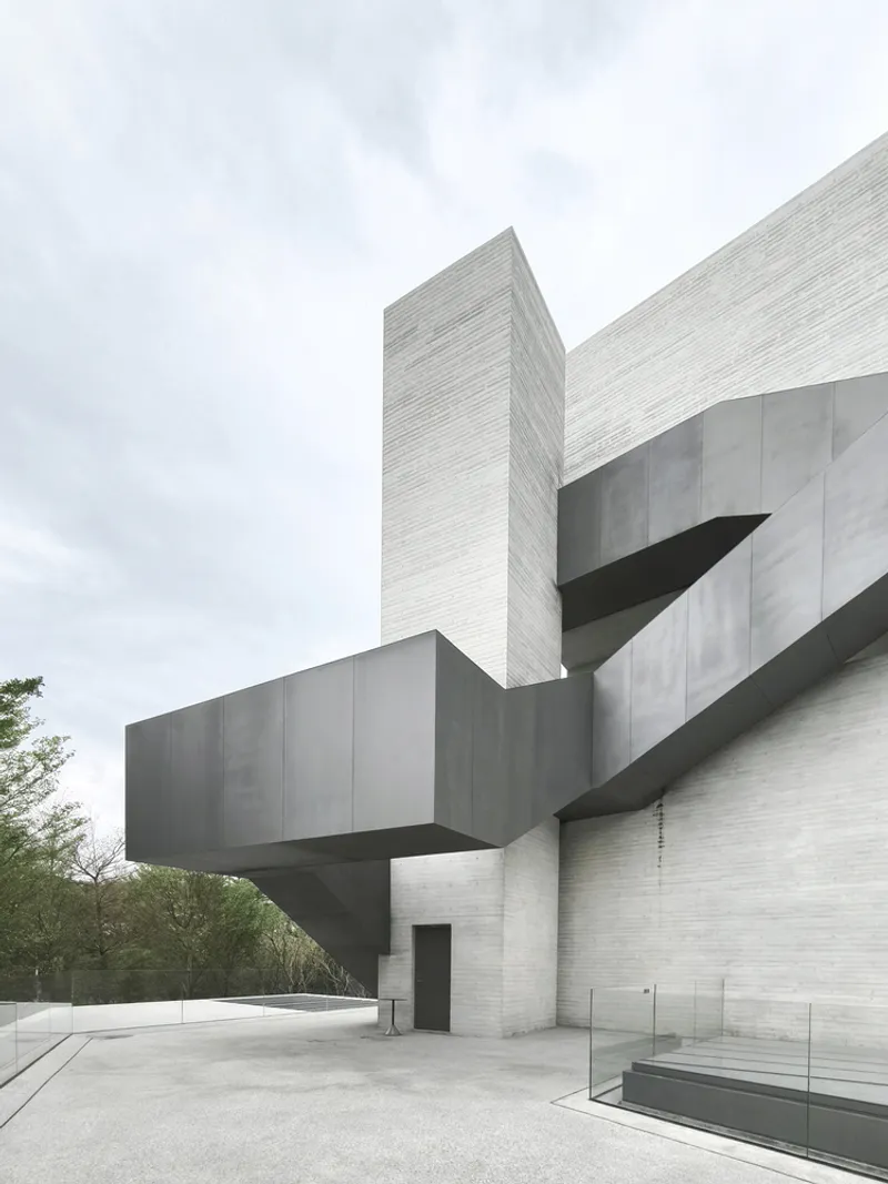 Cantilevered concrete volumes projecting from a textured white brick tower under an overcast sky