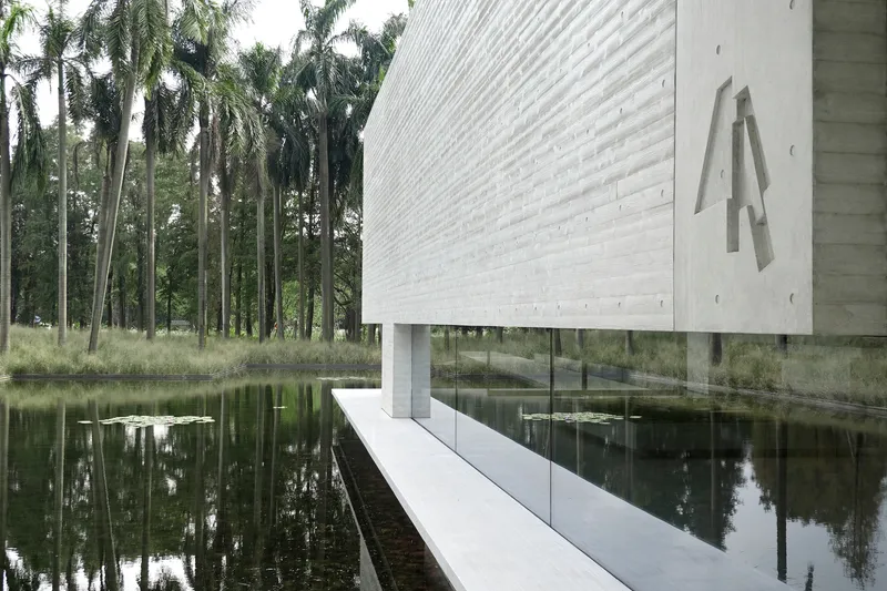 White painted brick volume on stilts extending over a still pond surrounded by palm trees