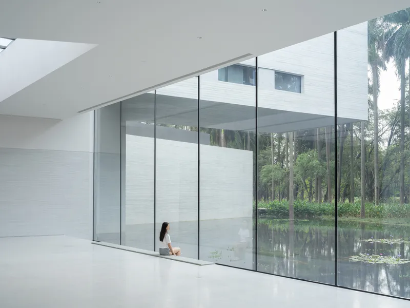 Floor-to-ceiling glazed wall reflecting a pond and trees with a seated figure in white interior