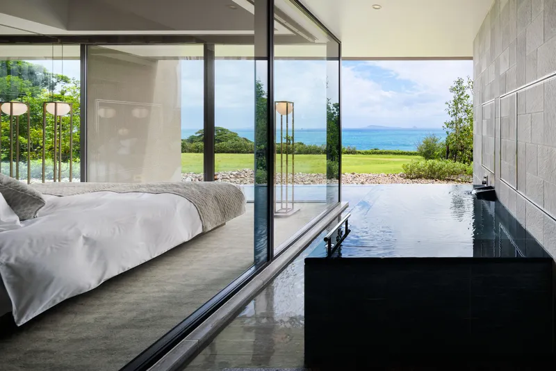 Bedroom with black stone bathtub threshold opening to glass walls overlooking lawn and distant water
