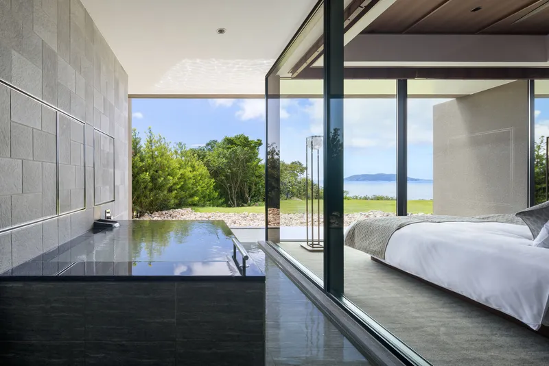 Black stone bathtub platform beside glazed wall opening to green lawn and lake under cloudy sky