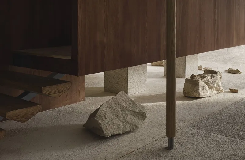 Detail of brass-trimmed walnut panel base with rough stone boulder casting shadow on terrazzo