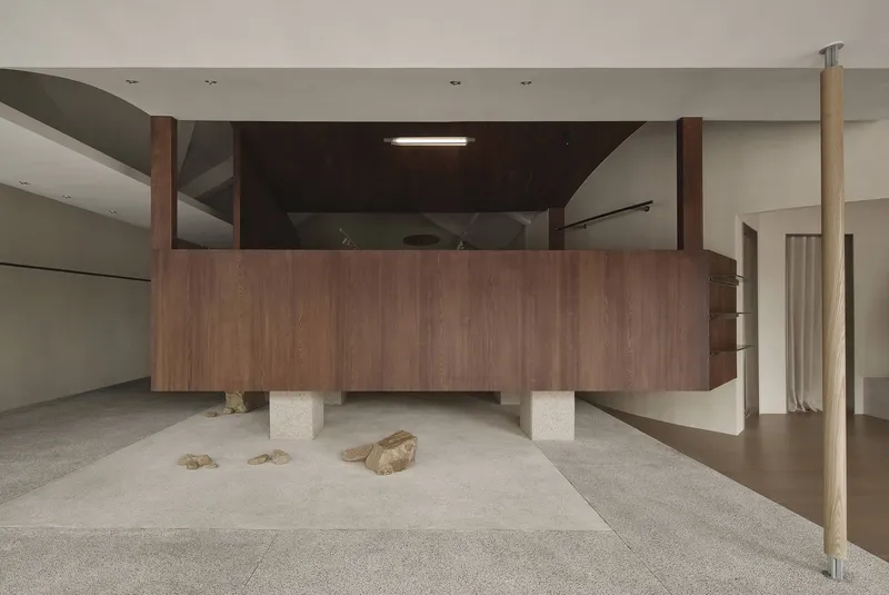 Walnut-clad volume elevated on square concrete blocks with loose stones scattered beneath on terrazzo