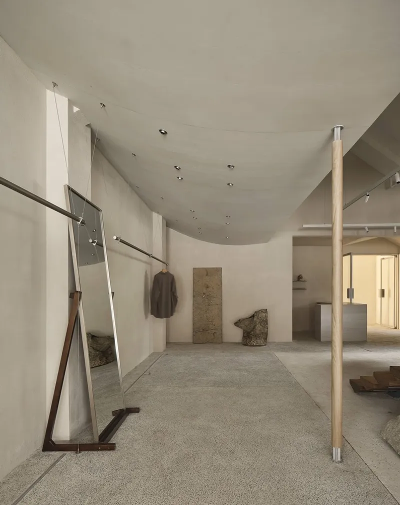 Gallery space with curved ceiling, timber column, leaning mirror, and hanging garment on terrazzo floor