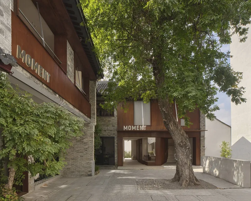 Facade showing corrugated metal panels and brick walls framed by a mature tree in the courtyard