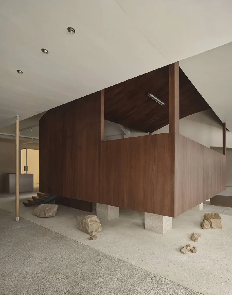 Freestanding walnut pavilion raised on concrete plinths with scattered rocks on terrazzo flooring