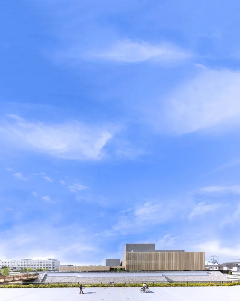 Long plaza view with pedestrians and cyclists before the horizontally slatted facade