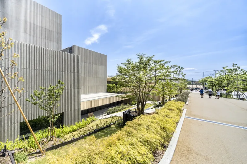 Landscaped walkway with planted beds along a ribbed concrete facade as visitors stroll in afternoon sun