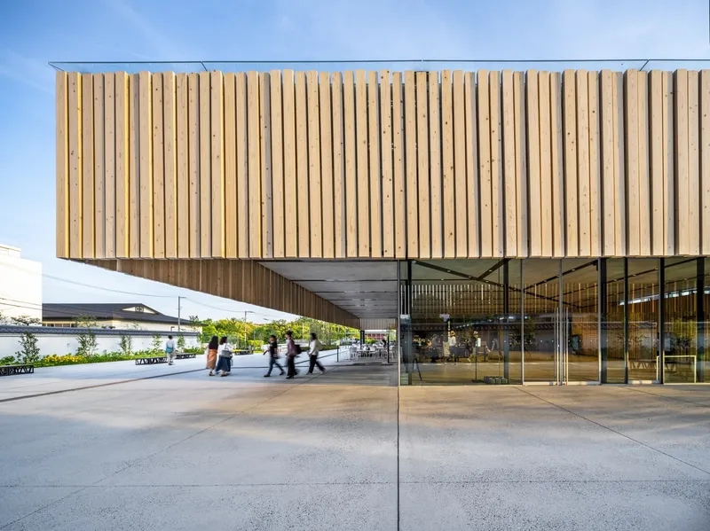 Cantilevered volume clad in vertical timber slats sheltering a covered plaza with glass walls and visitors below