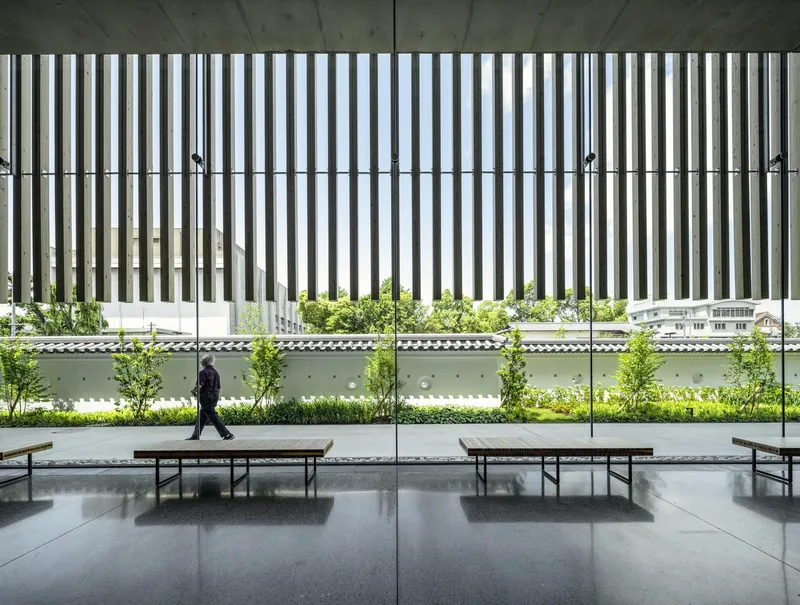 Covered outdoor terrace with vertical metal louvers above a planted bed with young trees and walking figure