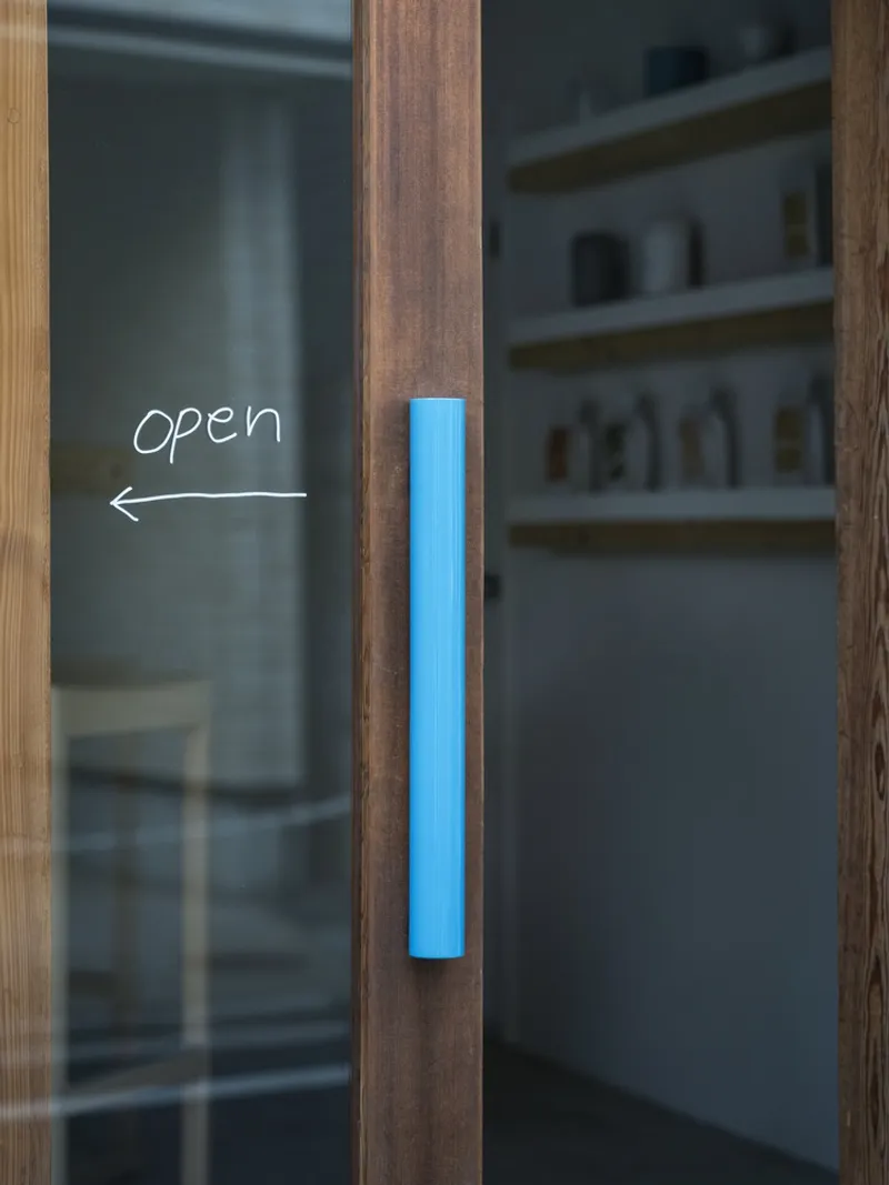 Glass entry door with timber frame and blue cylindrical handle beside handwritten open sign