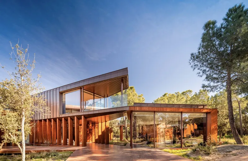 Two-story volume with corten cladding and glazed walls elevated on columns among pine trees at dusk