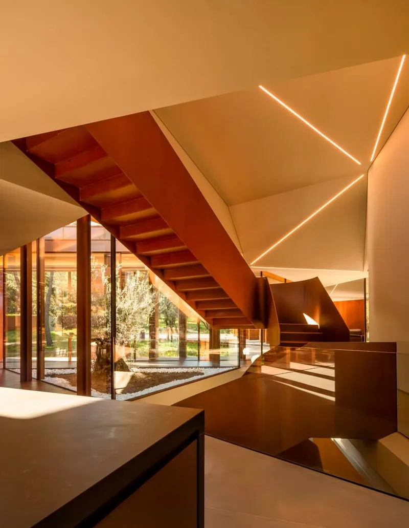 Interior staircase with steel stringers and linear ceiling lights beside a glazed courtyard with a tree