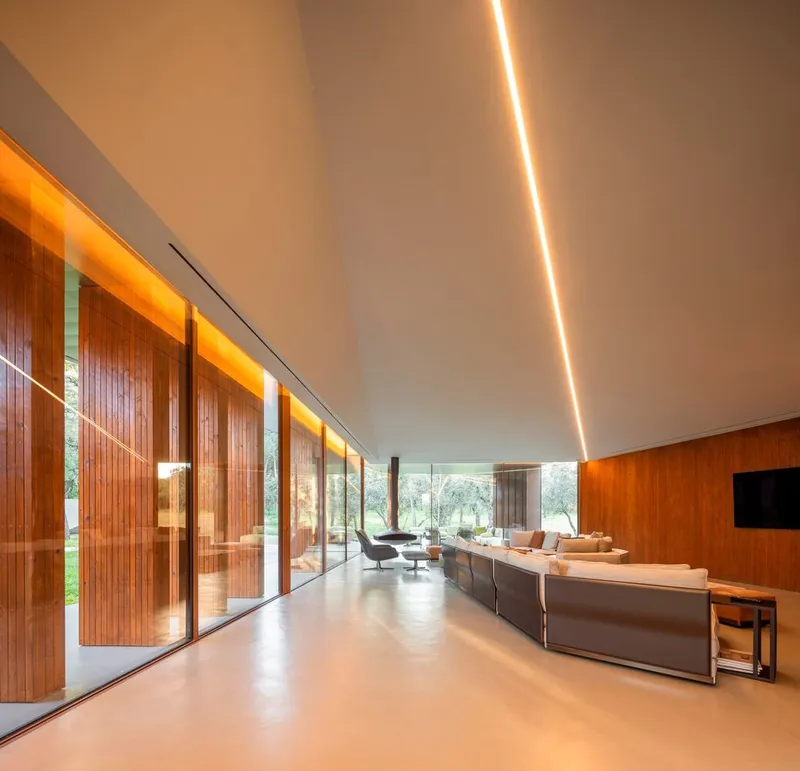 Interior living space with recessed ceiling lighting and floor-to-ceiling glazing overlooking olive trees at dusk