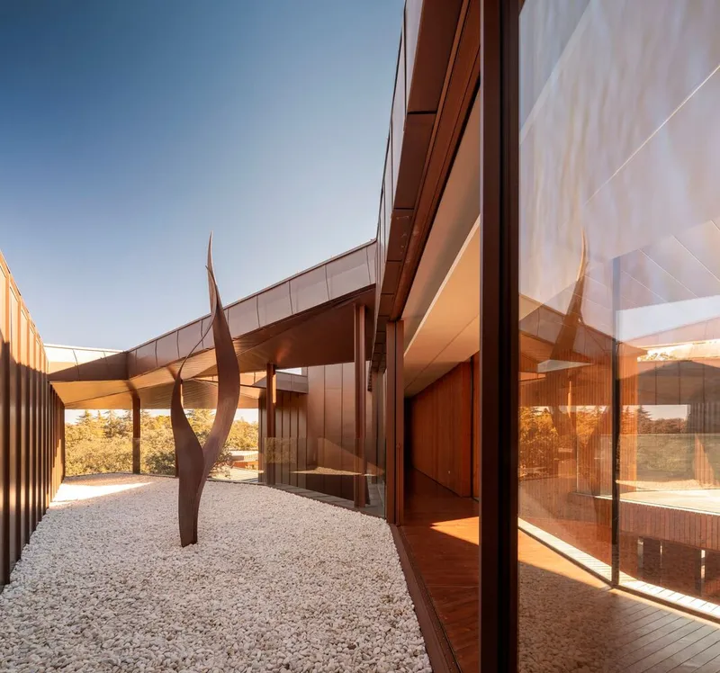 Gravel courtyard with cantilevered roof planes and a weathered steel sculpture beneath clear blue sky