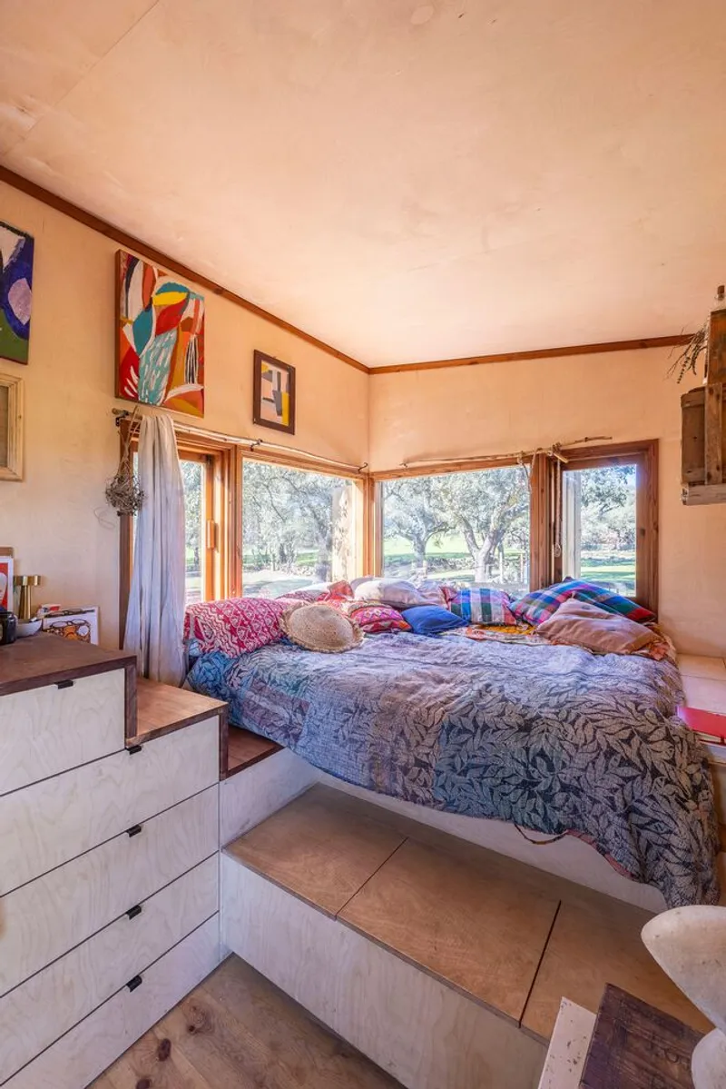 Built-in sleeping platform with corner windows framing oak grove in afternoon light
