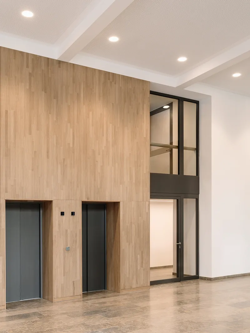 Entry lobby with vertical timber slat wall flanking two elevator doors and a glazed stairwell beyond