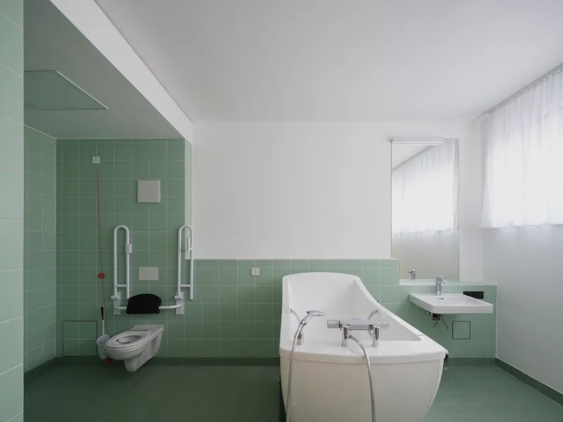 Accessible bathroom with mint green square tiles, wall-mounted toilet with support rails, and freestanding white bathtub