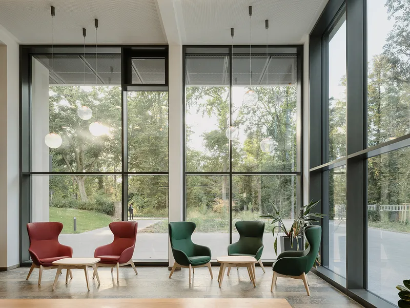 Double-height lounge with floor-to-ceiling steel-framed windows and colorful upholstered seating overlooking a green lawn