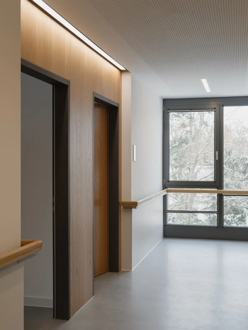 Corridor with wood-clad elevator surround, timber handrail, and dark-framed window overlooking bare winter trees