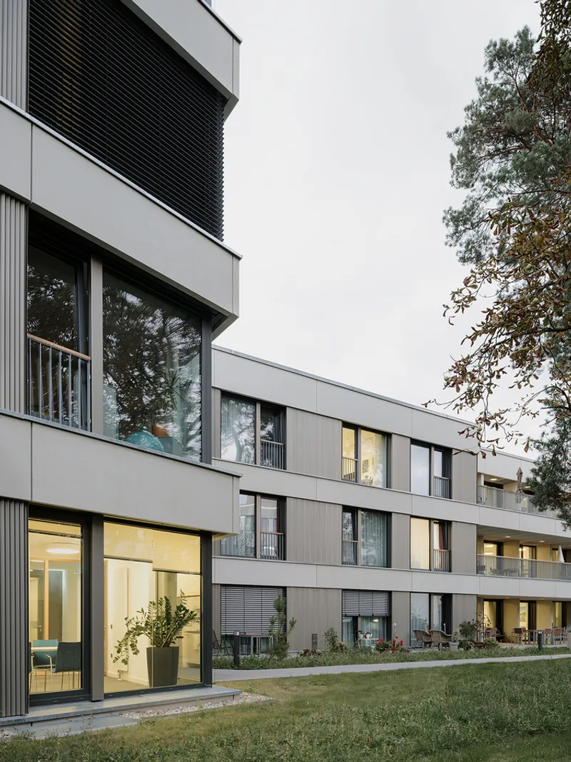 Stepped residential facade with grey panels and recessed balconies illuminated at dusk beside a lawn